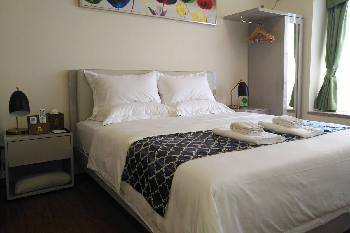 Une chambre d’hôtel avec un lit bien fait, une table de chevet avec une lampe et une télécommande, un tableau au mur et une fenêtre avec des rideaux.