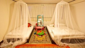 A room with two beds, each covered with a white canopy. There is a small table with a red tablecloth and a mirror above it. A colorful rug is placed in the center of the room. - Chezmoi Handicraft & HomeStay (Chiang Rai)