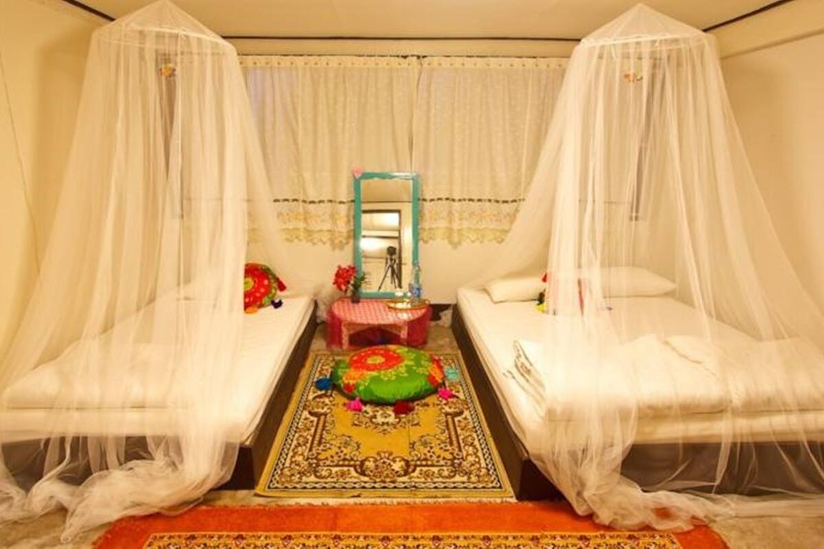 A room with two beds, each covered with a white canopy. There is a small table with a red tablecloth and a mirror above it. A colorful rug is placed in the center of the room.