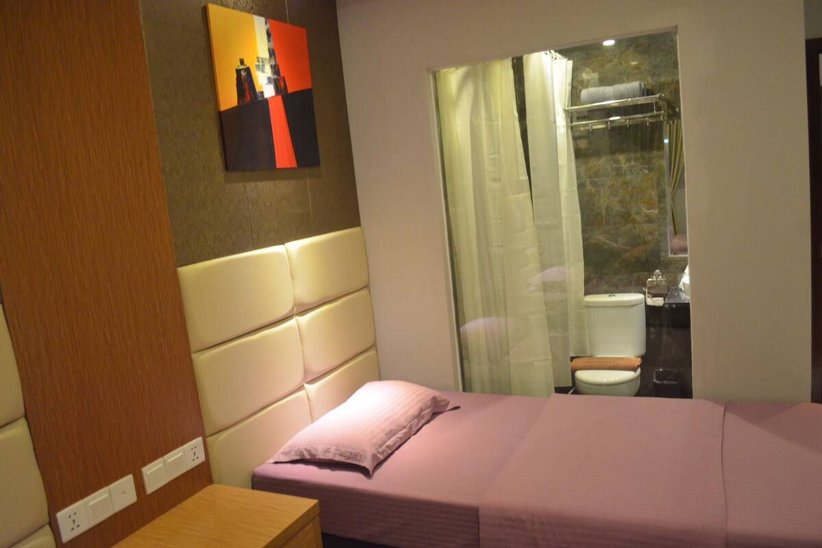 A hotel room with a bed, a bedside table, a bathroom visible through a glass partition, and a wall-mounted painting.