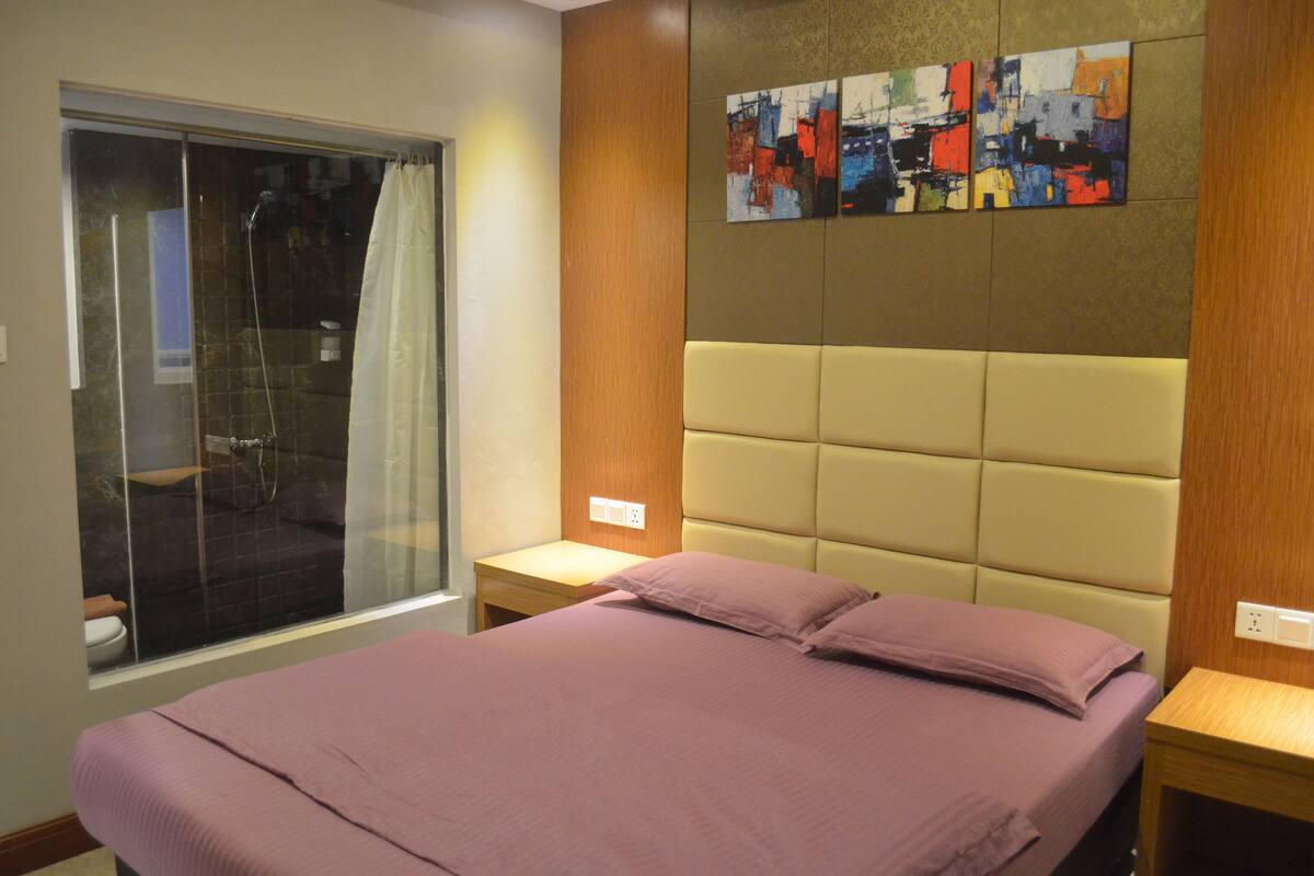 A bedroom with a bed, a window with a shower, and a wall with abstract art.