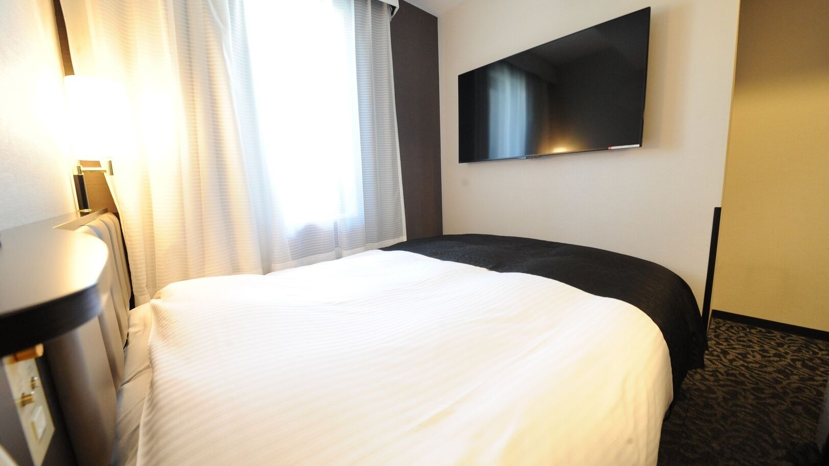 Una camera d'albergo con un letto, una TV a schermo piatto fissata al muro e una scrivania con una lampada.