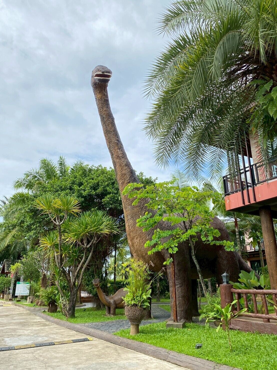 Una estatua de dinosaurio en un jardín exuberante con palmeras y un edificio al fondo.