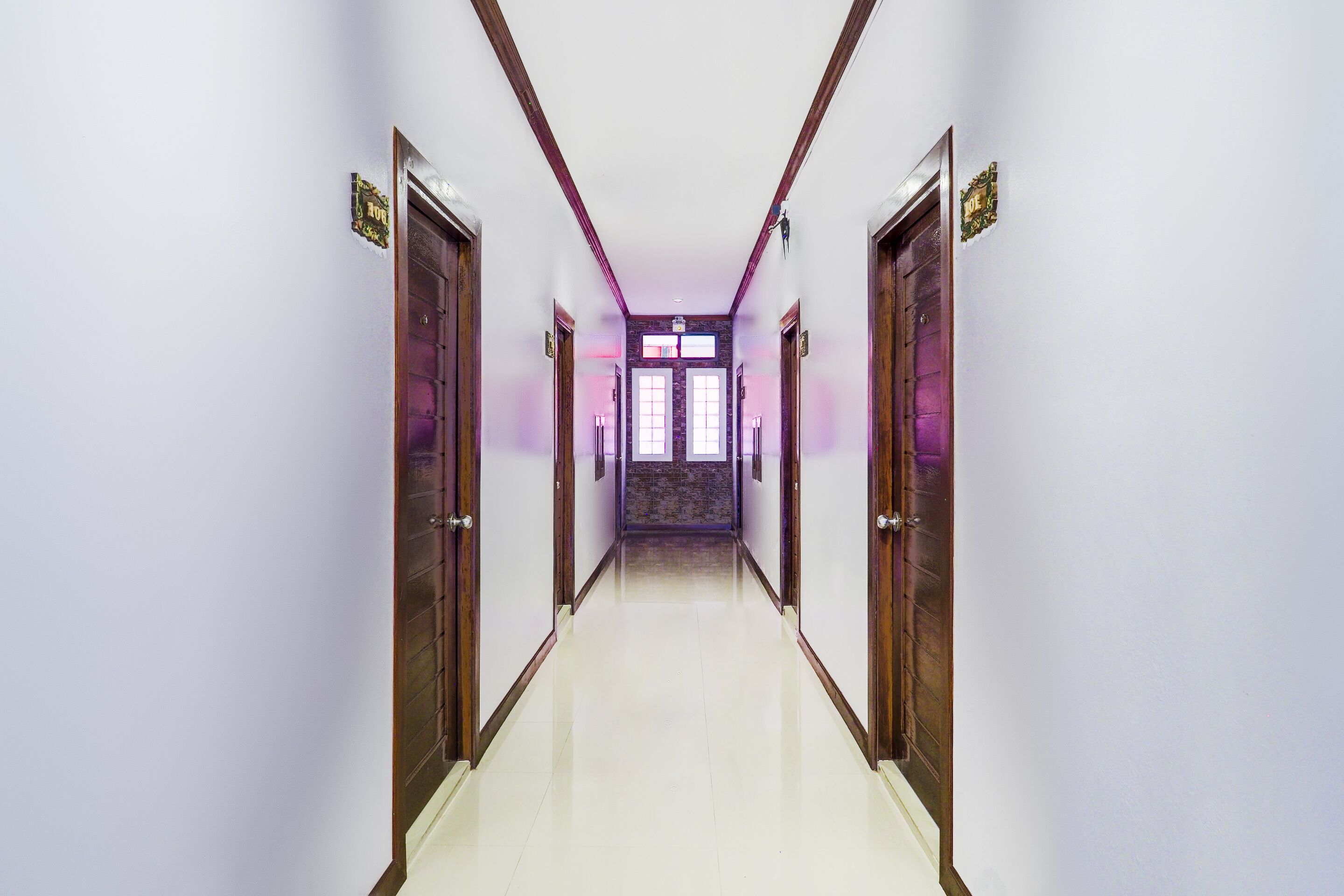 A long, narrow hallway with wooden doors on either side, leading to a room with a large window.