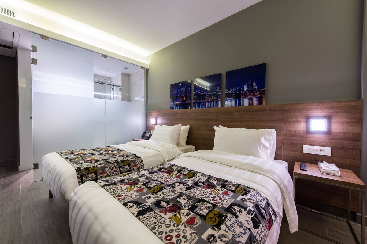 Habitación de hotel con dos camas, cabecera de madera, mesita de noche con un teléfono y un control remoto, y una pared con tres cuadros enmarcados.