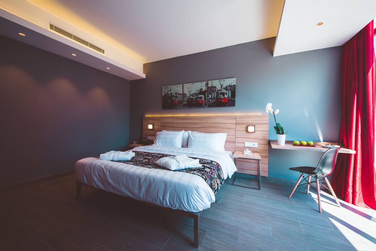 Una habitación de hotel moderna con una cama grande, un escritorio con una silla, una mesita con una planta y un cuadro de un paisaje urbano en la pared.