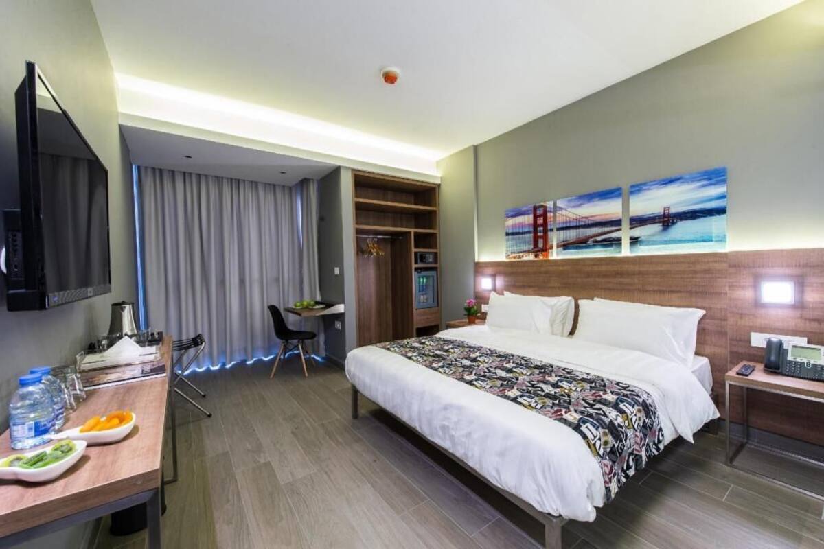 Una habitación de hotel con cama, escritorio, televisor, estantería y obras de arte en la pared.
