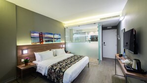 Habitación de hotel moderna con una cama grande, una mesita de noche con una planta, un panel divisor de vidrio, una televisión y una zona de kitchenette.