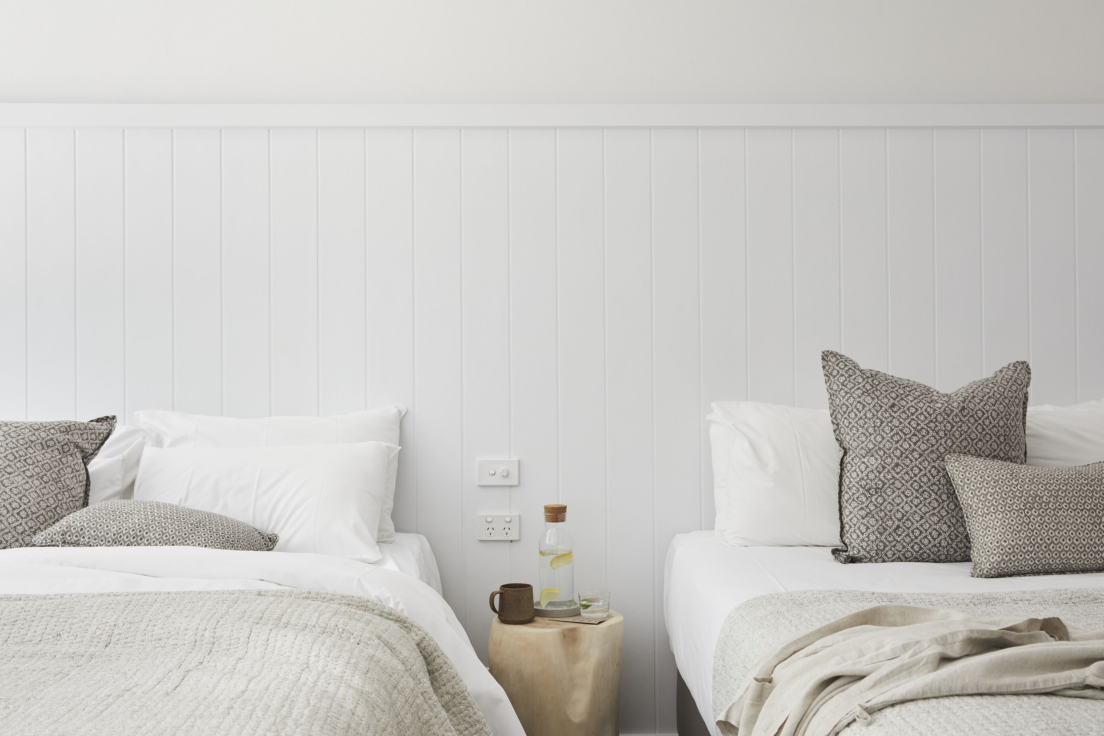 A bedroom with two beds, each with patterned pillows, a nightstand with a bottle and glass, and a white wall with a power outlet.