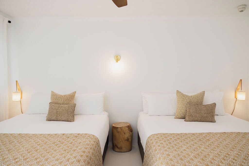 Une chambre d’hôtel avec deux lits, chacun recouvert d’une couvre-lit à motifs et agrémenté d’oreillers assortis. Un petit tabouret en bois est placé entre les lits, et un luminaire mural est fixé au-dessus du centre de la pièce.