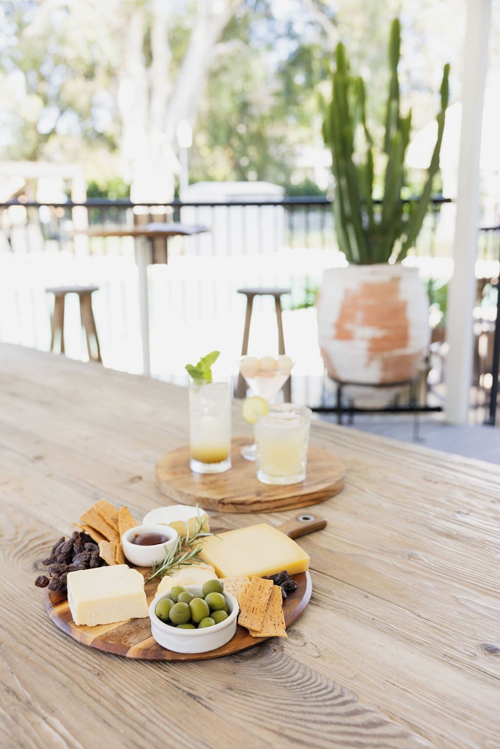 A wooden table with a cheese board, olives, crackers, and drinks.