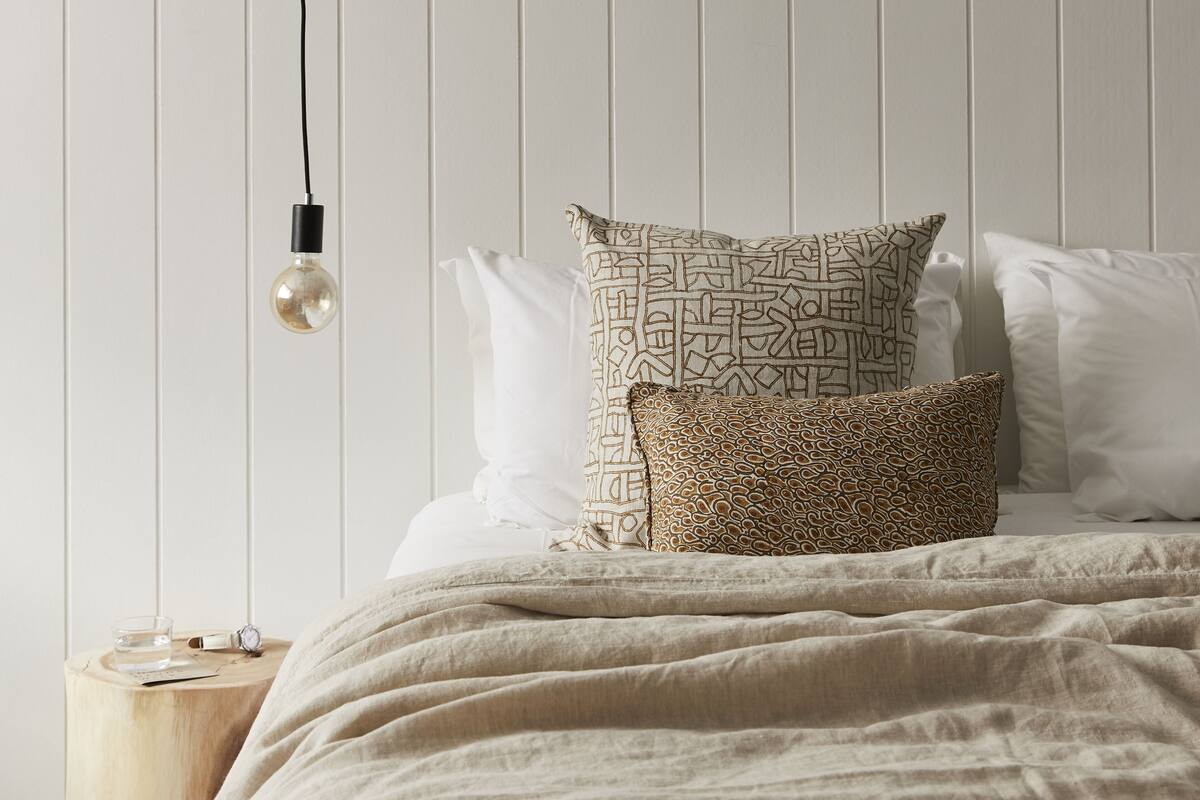 A neatly made bed with patterned pillows, a bedside table with a glass of water, and a hanging light bulb.