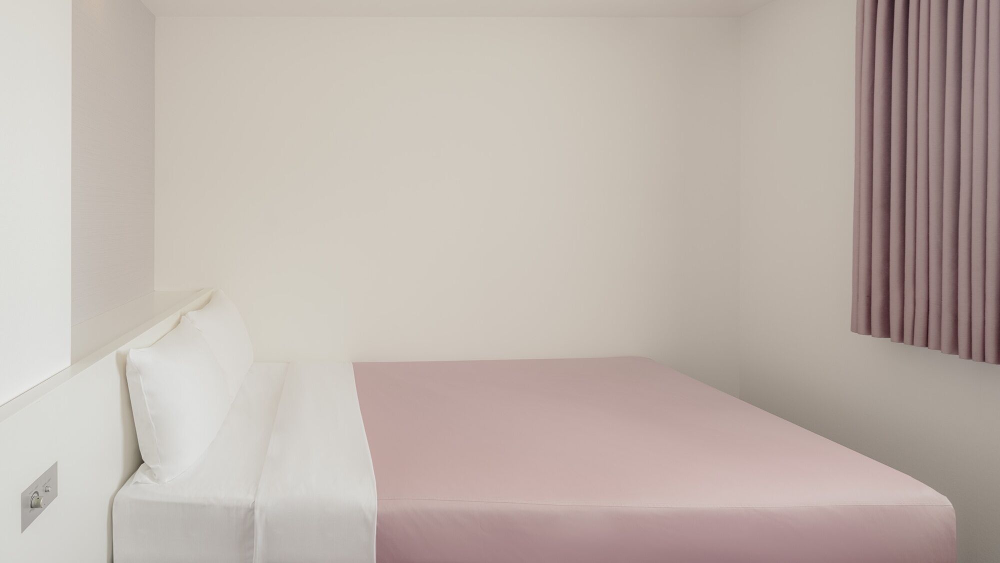 Habitación pequeña con una cama individual, cabecera blanca, colchón rosa y cortina instalada en la pared.