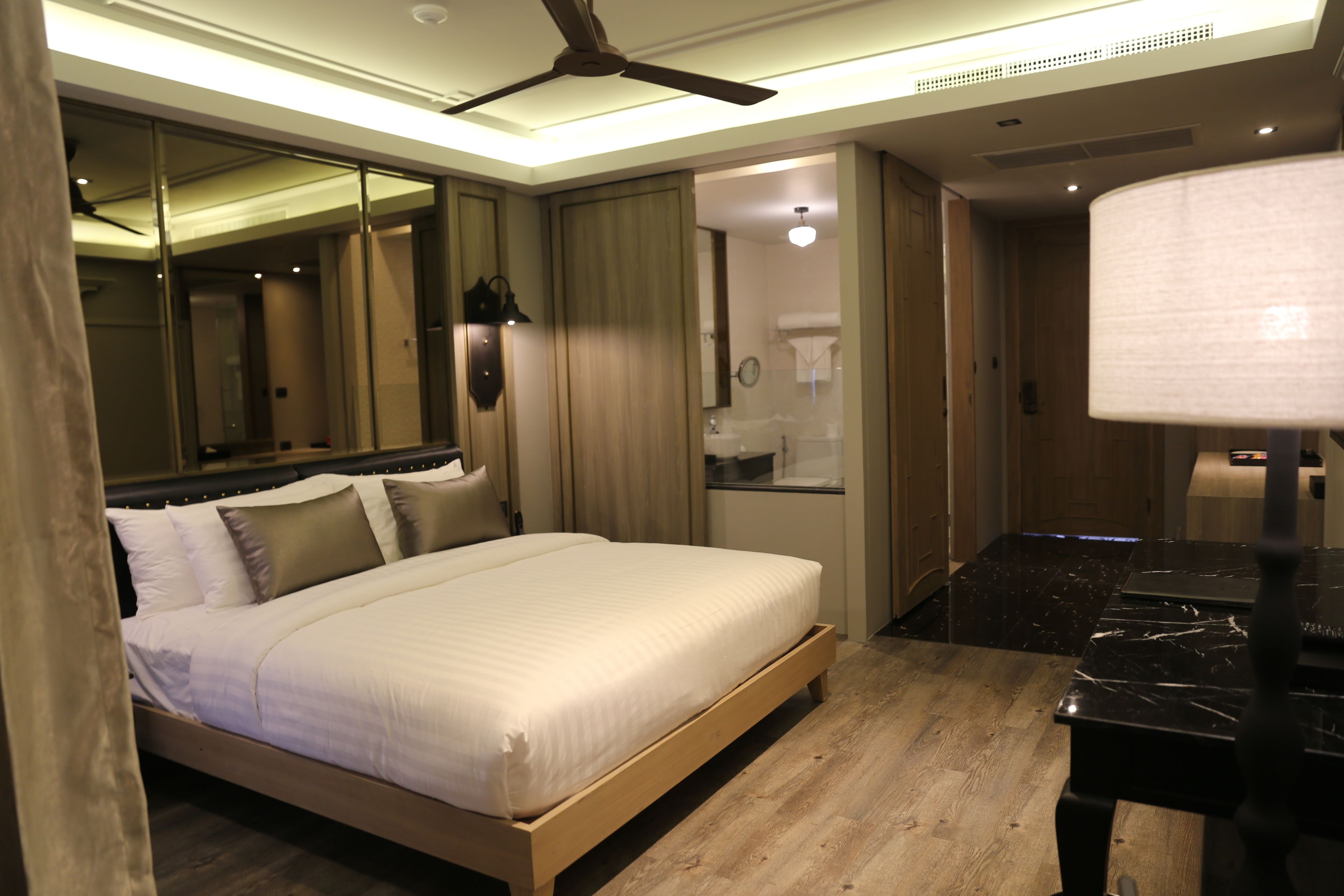 A modern hotel room with a wooden bed, a desk, a mirror, and a bathroom visible through an open door.