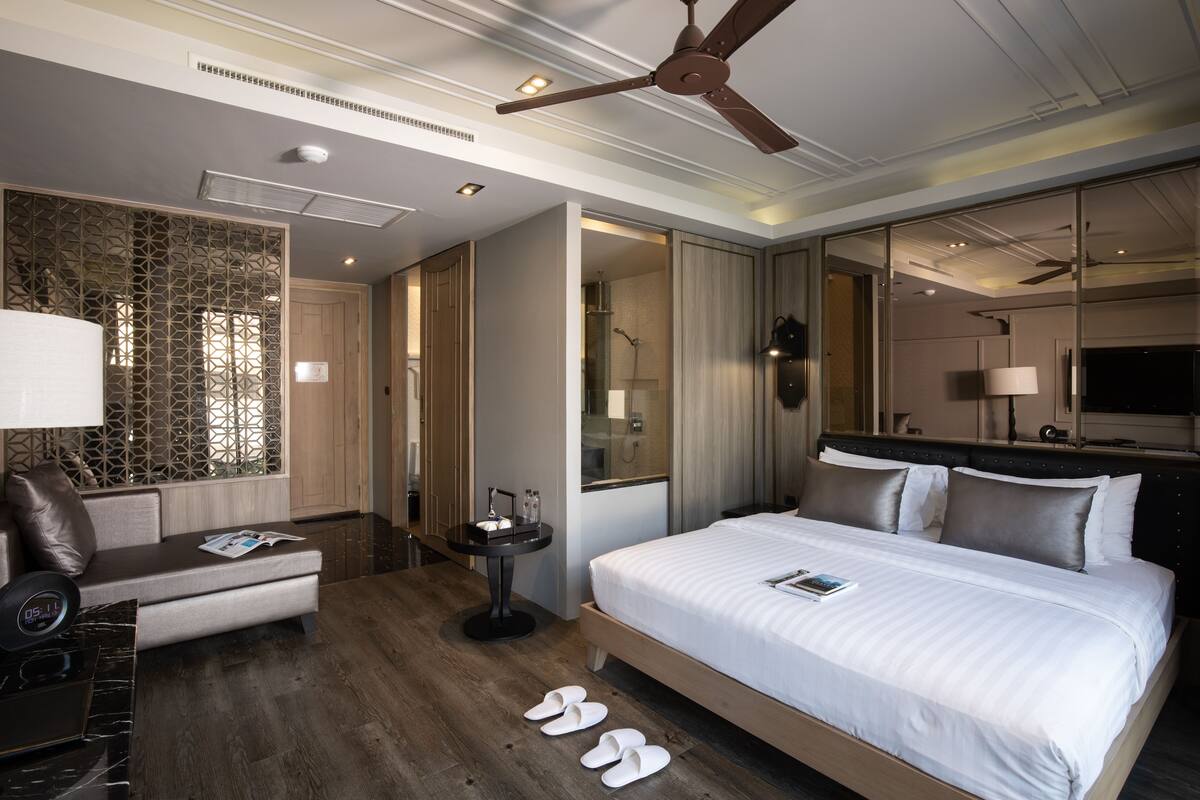 Ein modernes Hotelzimmer mit einem großen Bett, einer Sitzecke mit Sofa und Sesseln, einem kleinen Tisch mit Lampe und einem Deckenventilator.