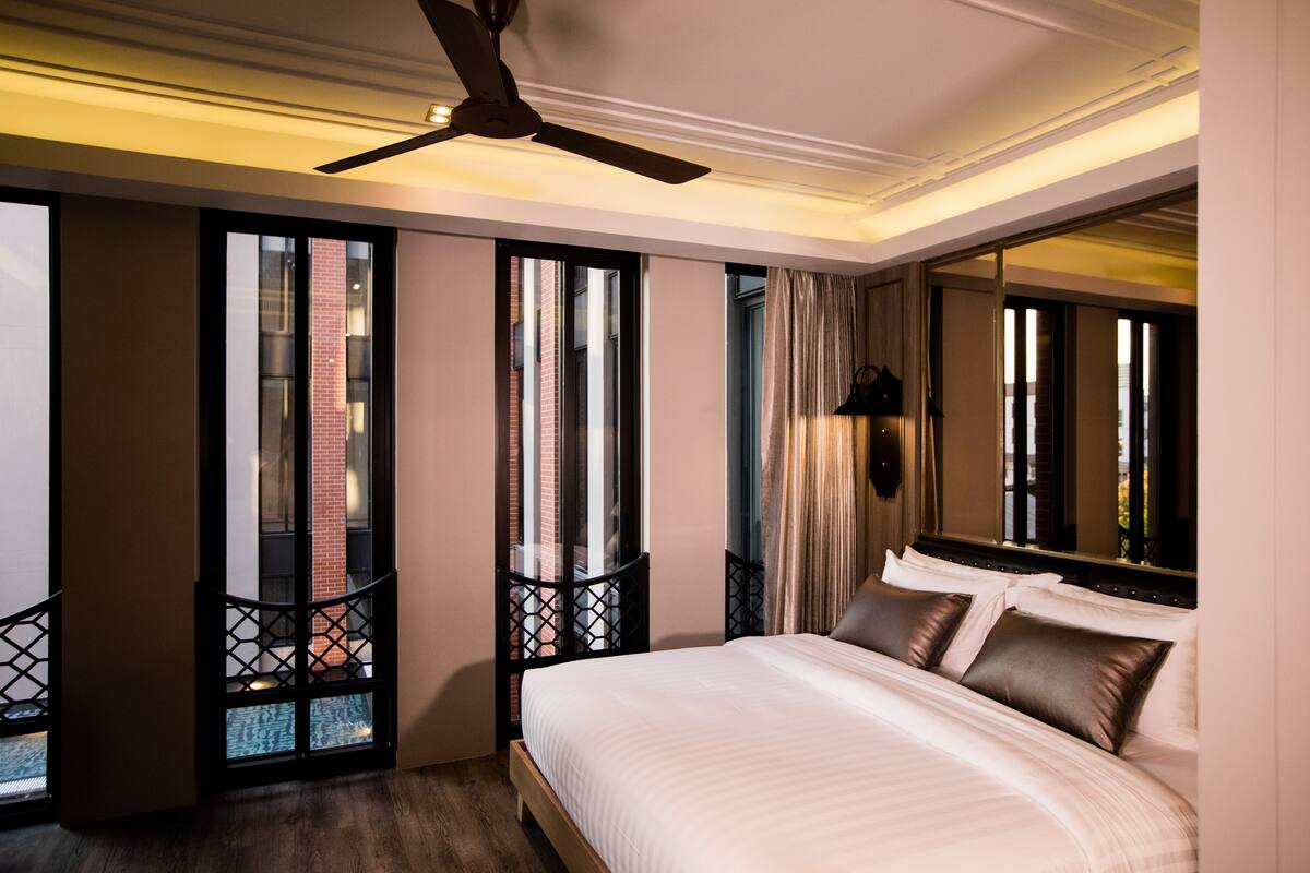 Ein Hotelzimmer mit einem großen Bett, einem Deckenventilator und Blick auf ein Ziegelgebäude durch die Fenster.