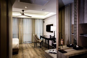 A hotel room with a bed, a desk, a chair, a standing lamp, and a large window with curtains.
