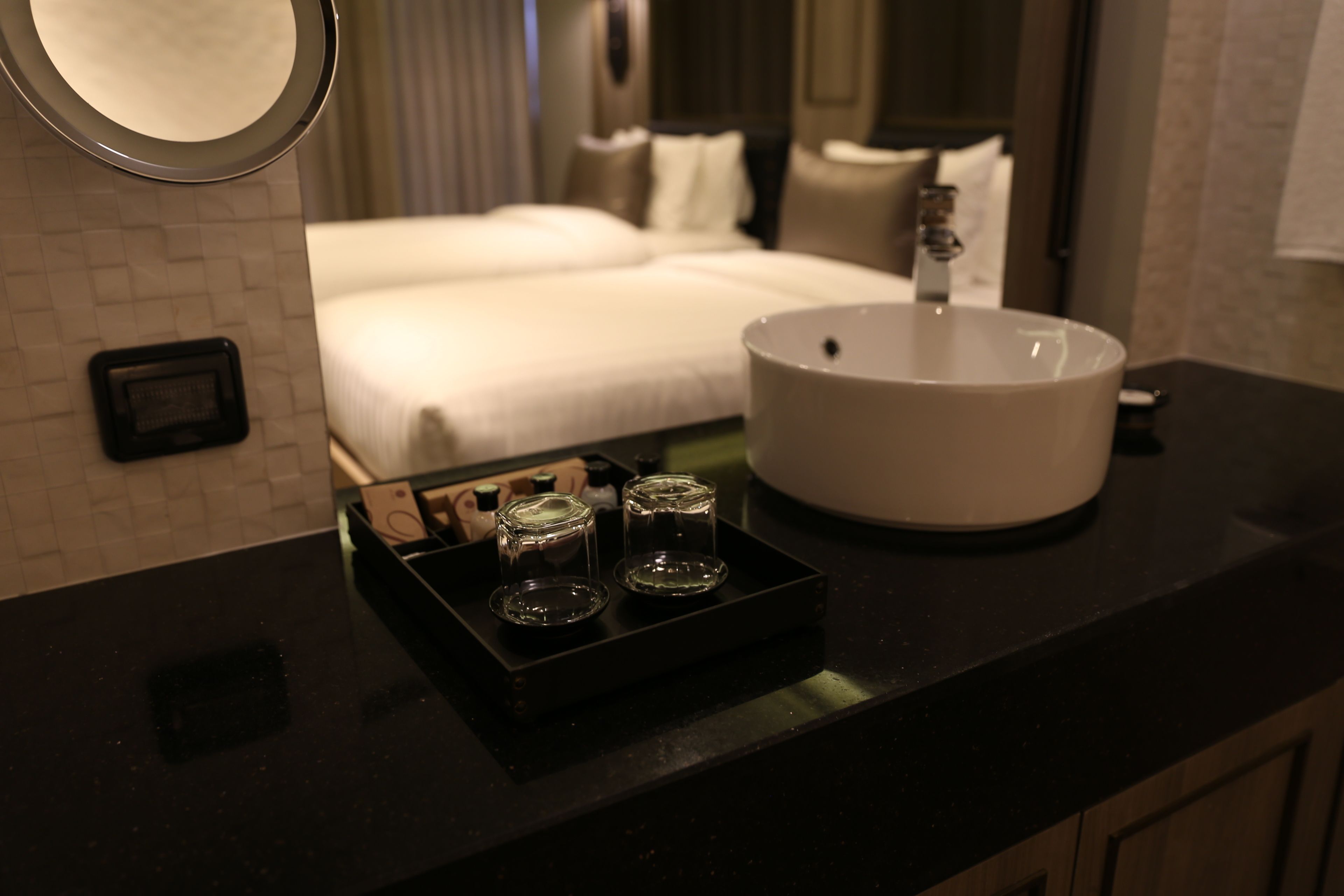 Un baño de hotel con encimera negra, un lavamanos blanco y dos vasos de vidrio sobre una bandeja.