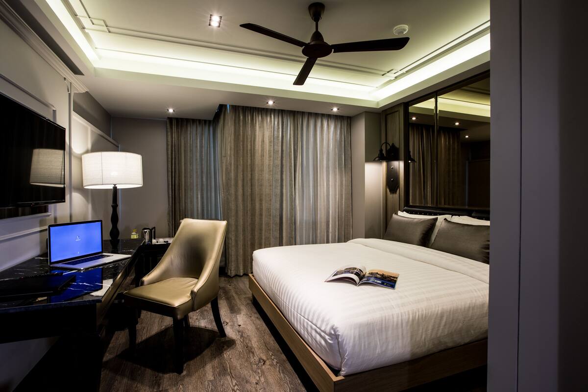 Ein Hotelzimmer mit einem Bett, einem Schreibtisch mit Laptop, einem Sessel und einem Deckenventilator, sowie einem großen Spiegel.