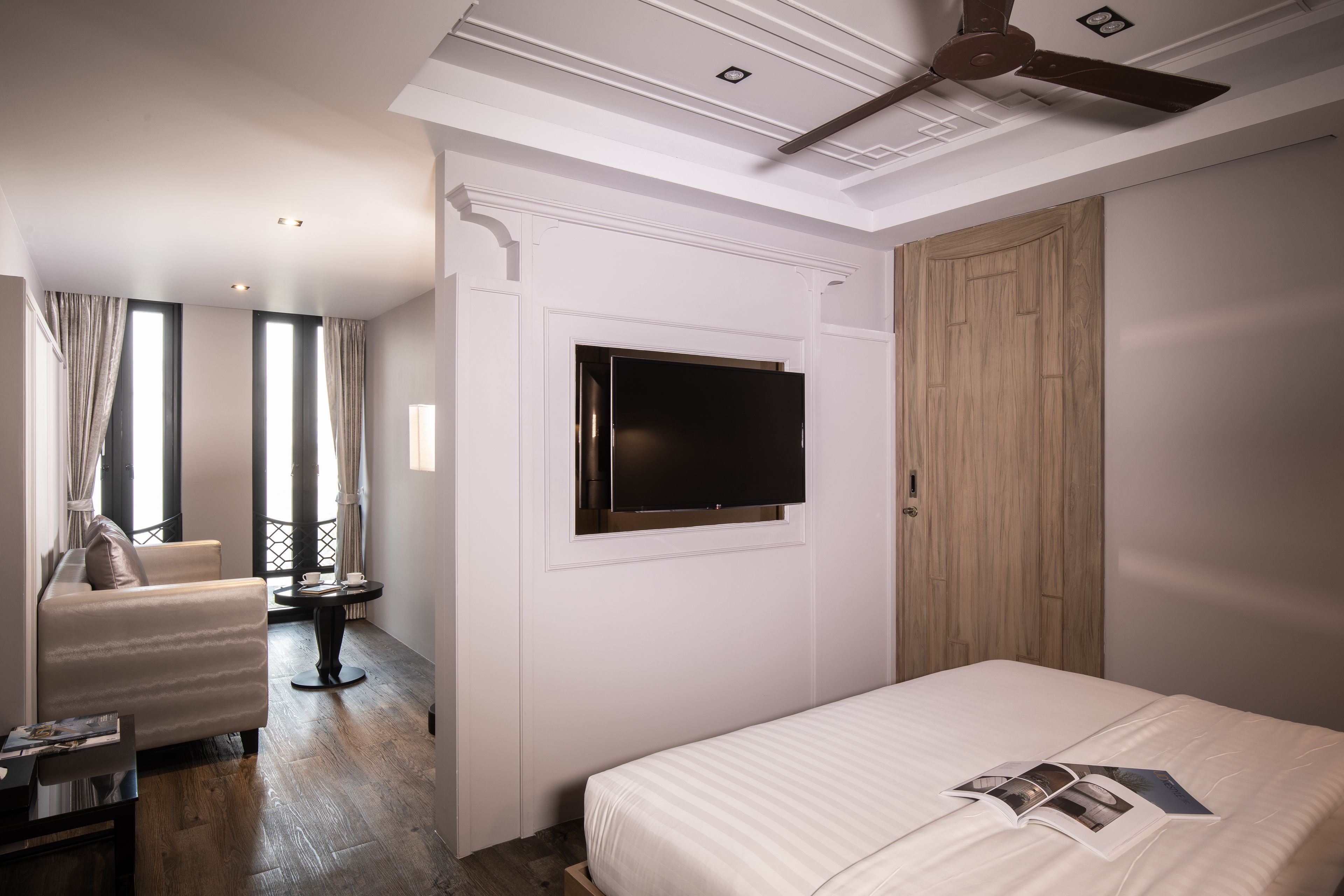 Una habitación de hotel moderna con una cama, un televisor montado en la pared, un armario de madera, una zona de estar con una silla y una mesa, y ventanas grandes con cortinas.