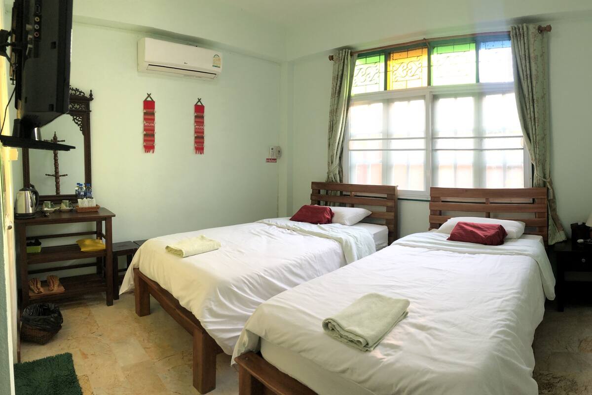 A room with two beds, a wooden nightstand, a table with a kettle, and a window with curtains.