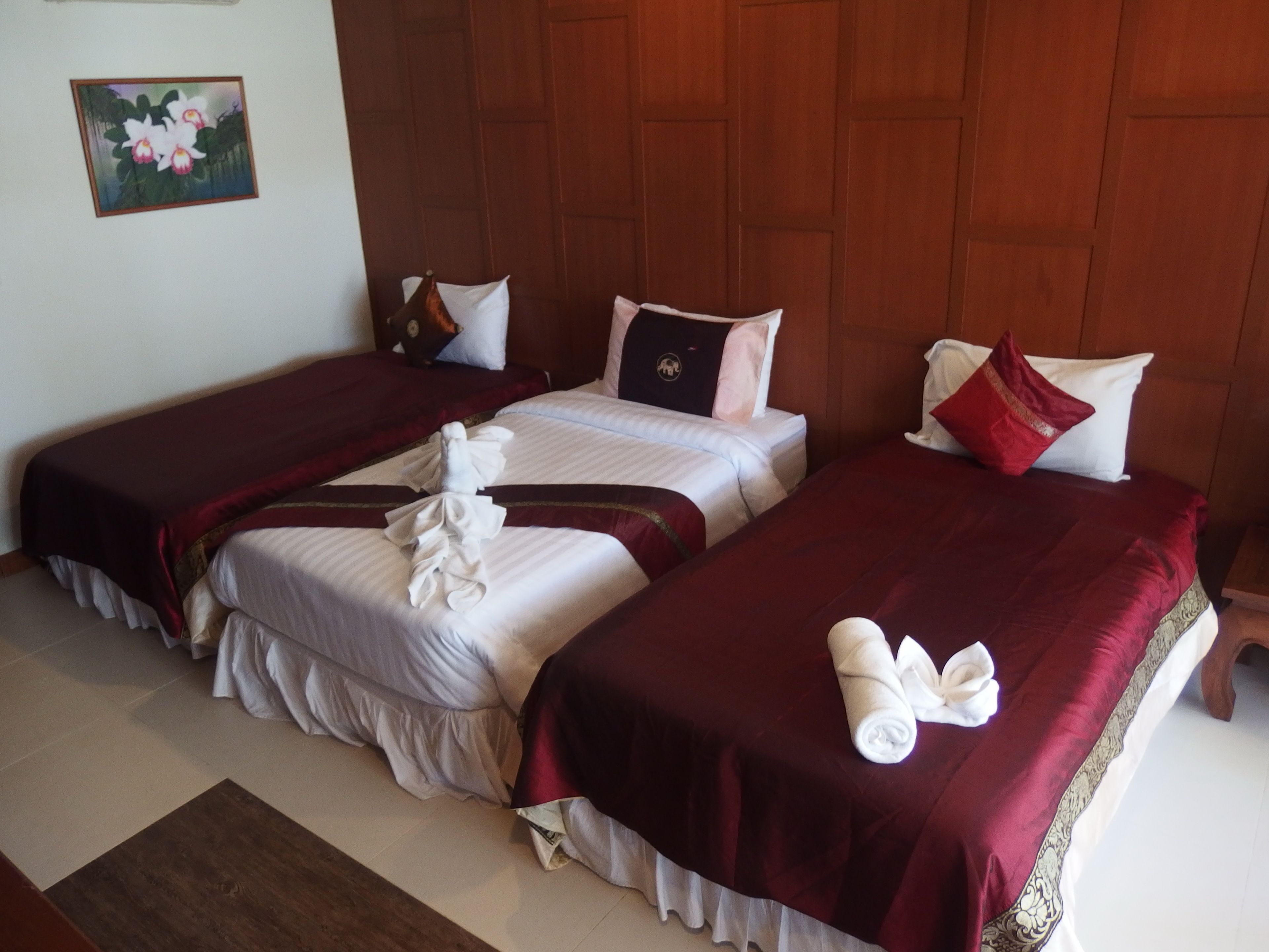 A hotel room with three beds, each with a red bedspread and white linens. There are towels folded on the beds, and a painting of flowers on the wall.
