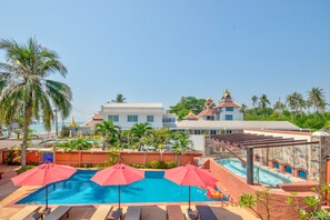 A swimming pool with red umbrellas, lounge chairs, and a view of a building with a golden temple-like structure in the background. - Palm Beach Resort (Pranburi)