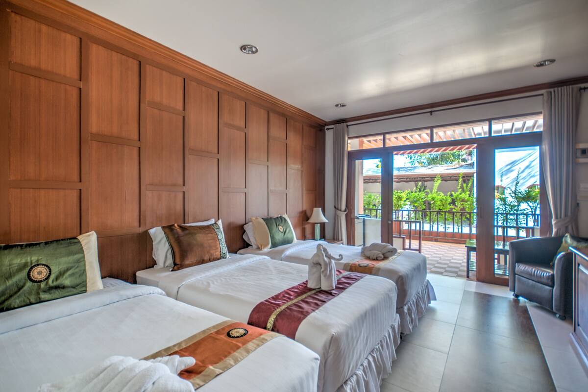 Une chambre d’hôtel avec deux lits, un lambris en bois, un balcon donnant sur un espace verdoyant et un coin salon avec un canapé et des fauteuils.