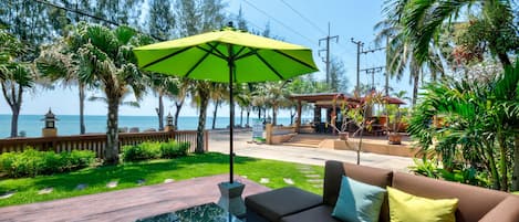 A patio with a glass table, a brown sofa with cushions, a green umbrella, and a view of the sea and palm trees.