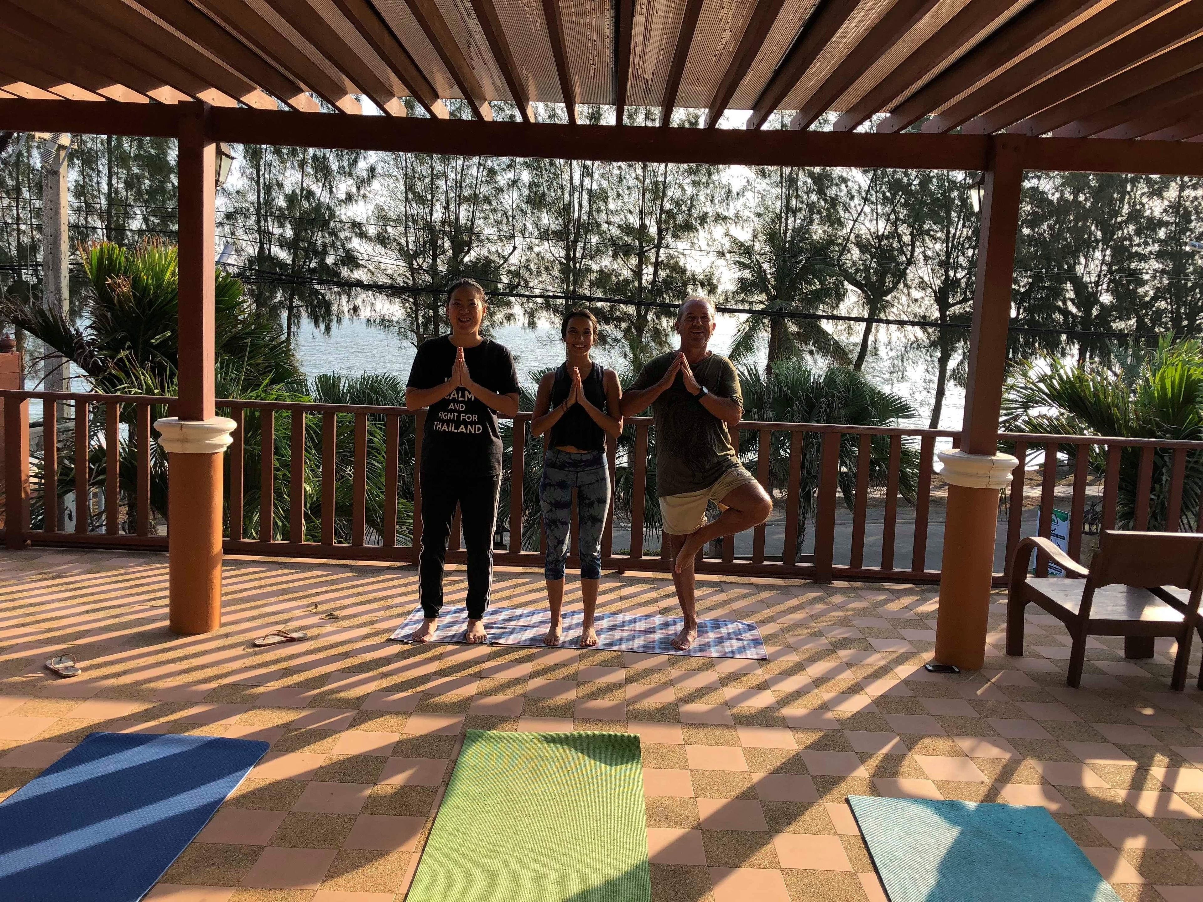 Tre personer utövar yoga på mattor under en träpergola med utsikt över träd och en vattenyta.