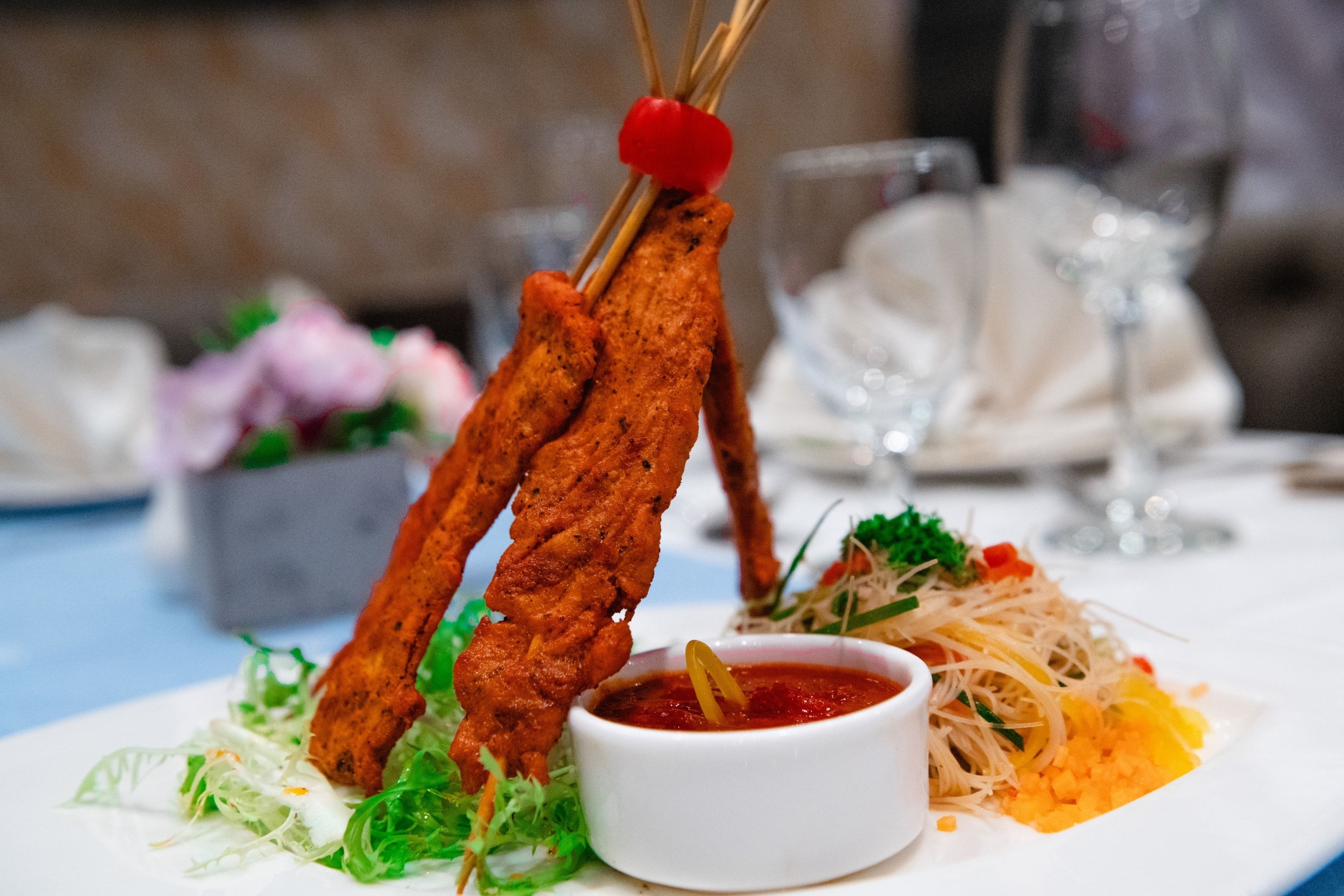 A plate with two skewers of grilled meat, a bowl of sauce, and a side of noodles.