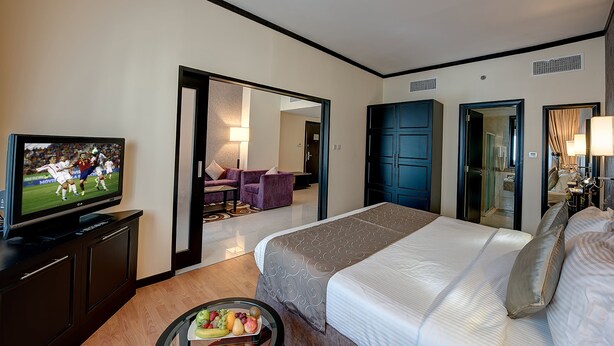A hotel room with a bed, a TV, a fruit bowl, and a view of the hallway. - Grandeur Hotel (Dubai)