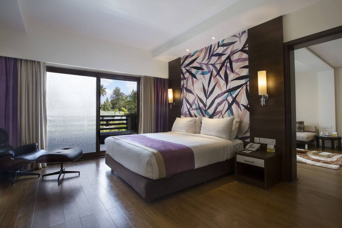 Quarto de hotel moderno com uma cama grande, uma escrivaninha com cadeira, uma mesa de cabeceira com luminária, e vista para o exterior através de uma porta de correr.
