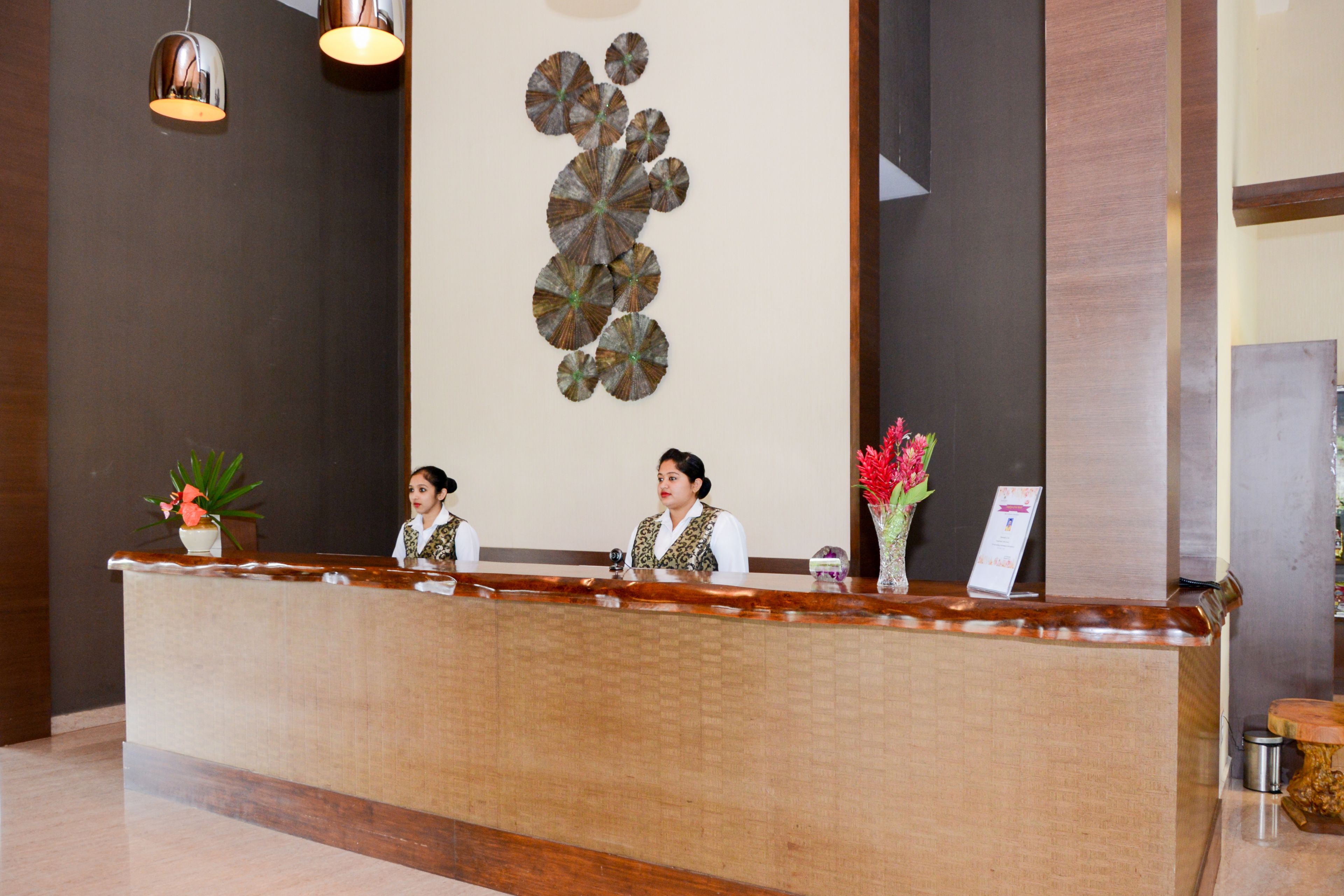 A reception desk with two staff members, a decorative wall, and a vase with flowers.