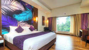 A hotel room with a large bed, a desk, a chair, a window with curtains, and a wall with a leaf pattern. - Purple Palms Resort & Spa (Somvarpet)
