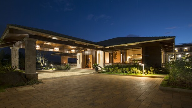 A modern building with a covered entrance, stone pillars, and a well-lit garden area. - Purple Palms Resort & Spa (Somvarpet)