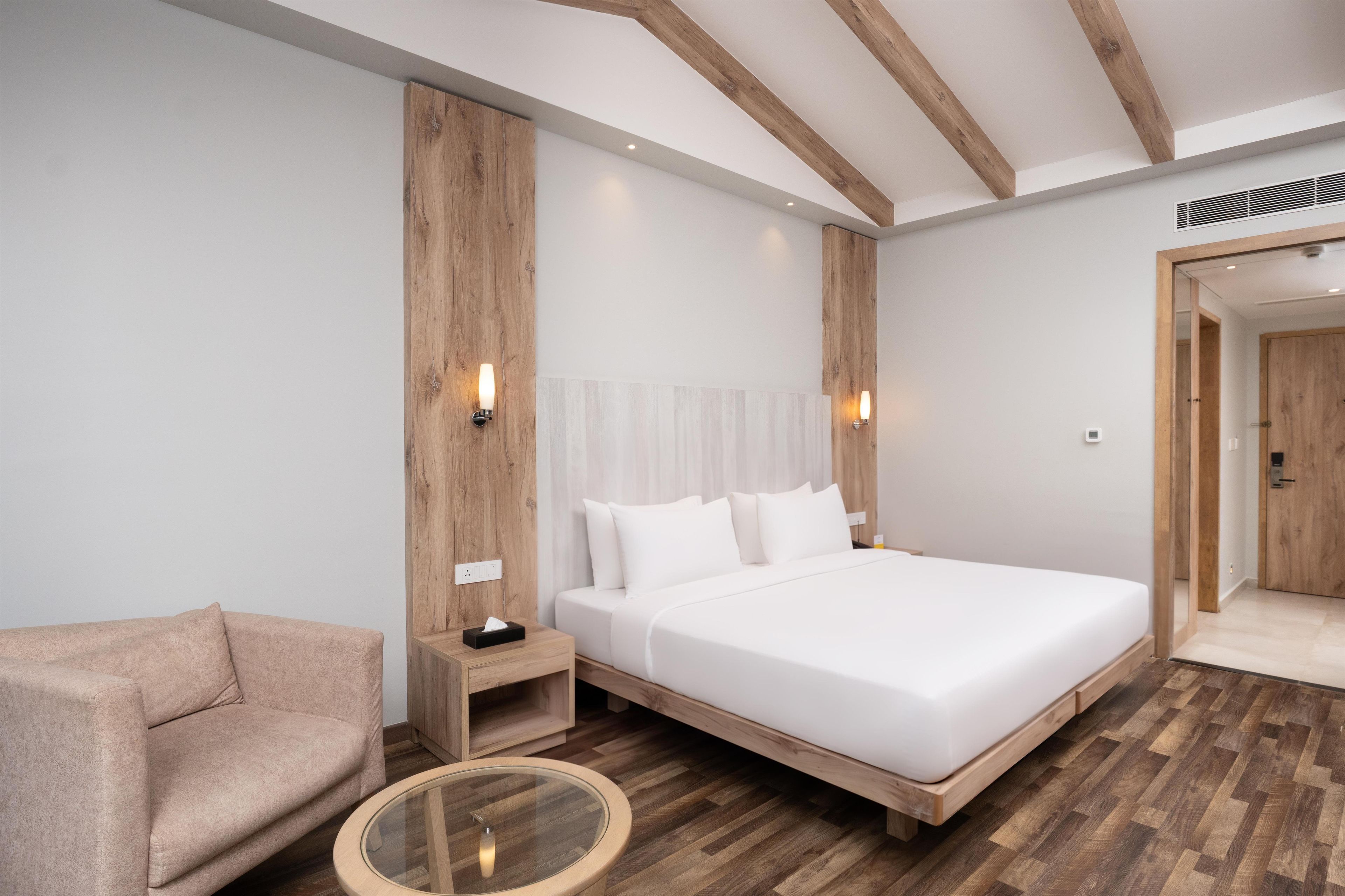 A modern hotel room with a large bed, a wooden nightstand, a round glass table, and a beige armchair.