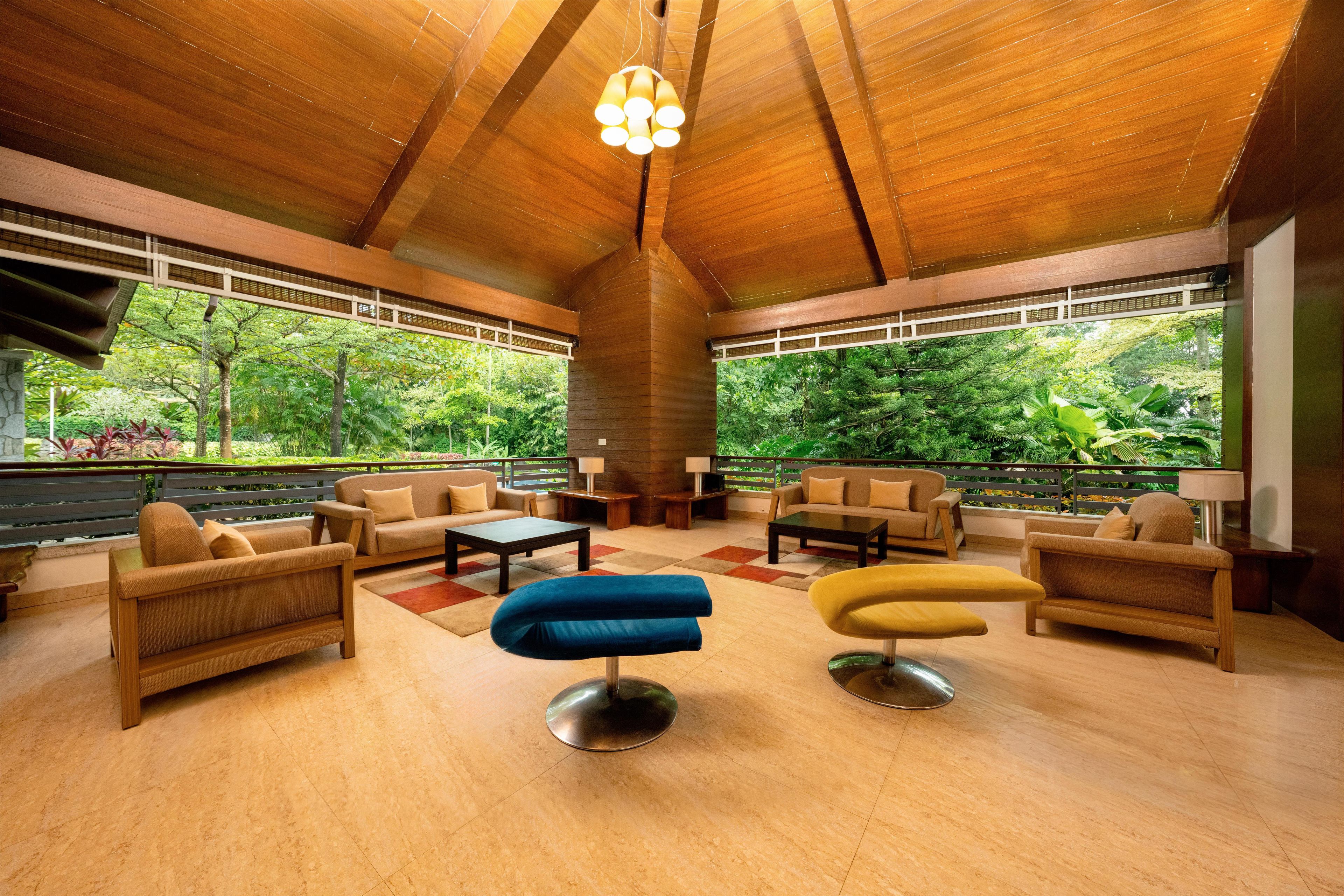 A spacious lounge area with a high ceiling, wooden beams, and large windows offering a view of lush greenery.