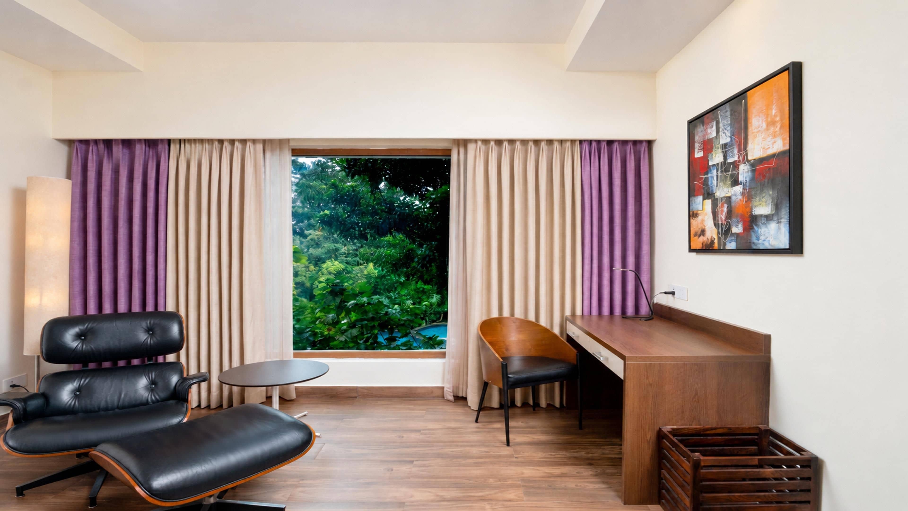 A room with a desk, two chairs, a small table, and a view of greenery outside.