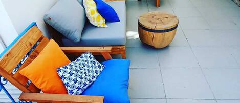 A sunlit outdoor seating area with wooden lounge chairs and sofas, a round wooden table, and a potted plant.