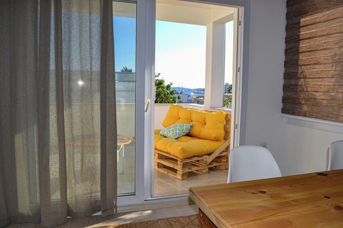 Una habitación con una puerta corrediza de vidrio que da a un balcón con zona de estar acolchada de color amarillo y una mesa de madera.