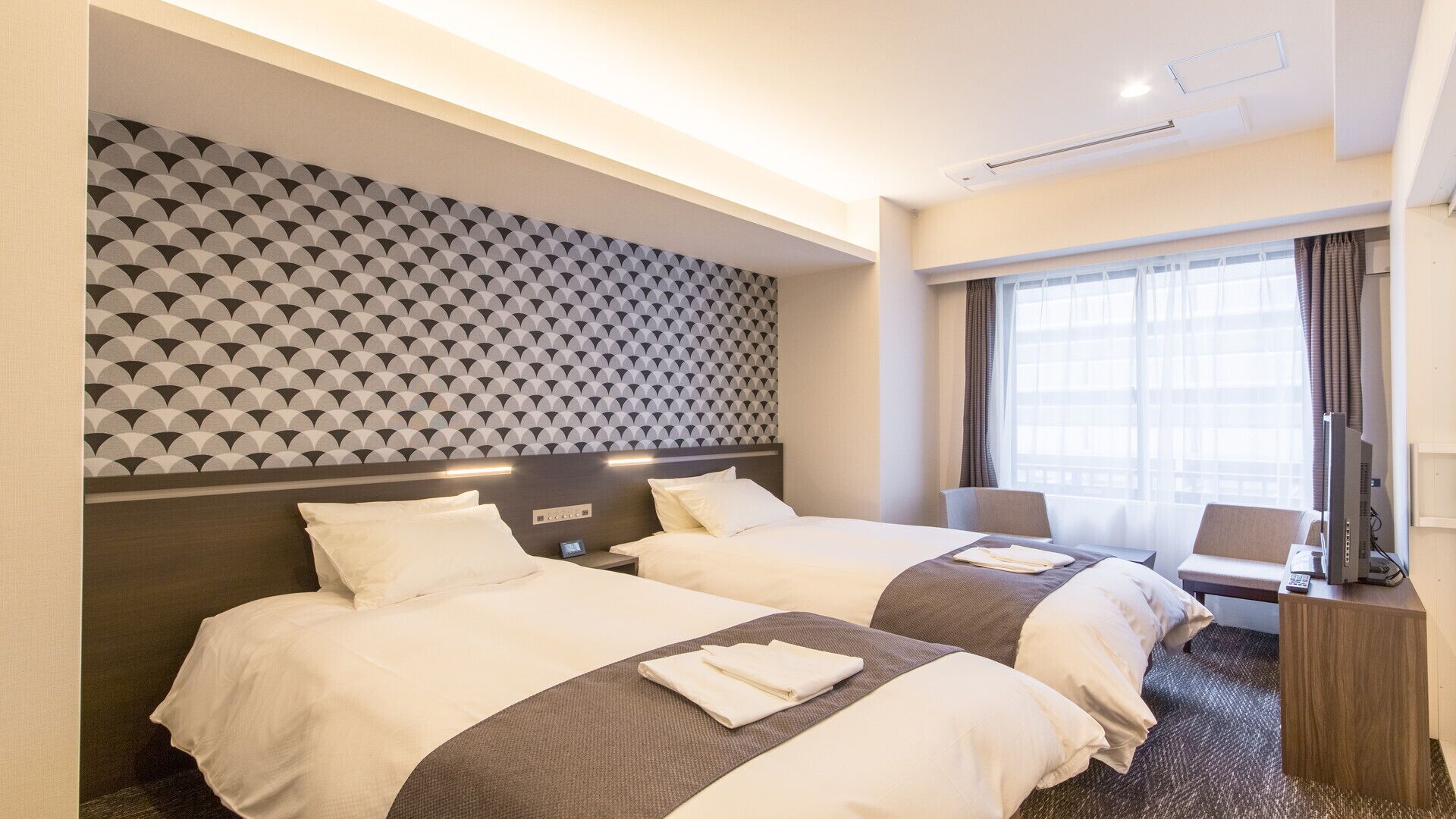 Habitación de hotel con dos camas, un papel tapiz estampado, una ventana con cortinas y un escritorio con ordenador.