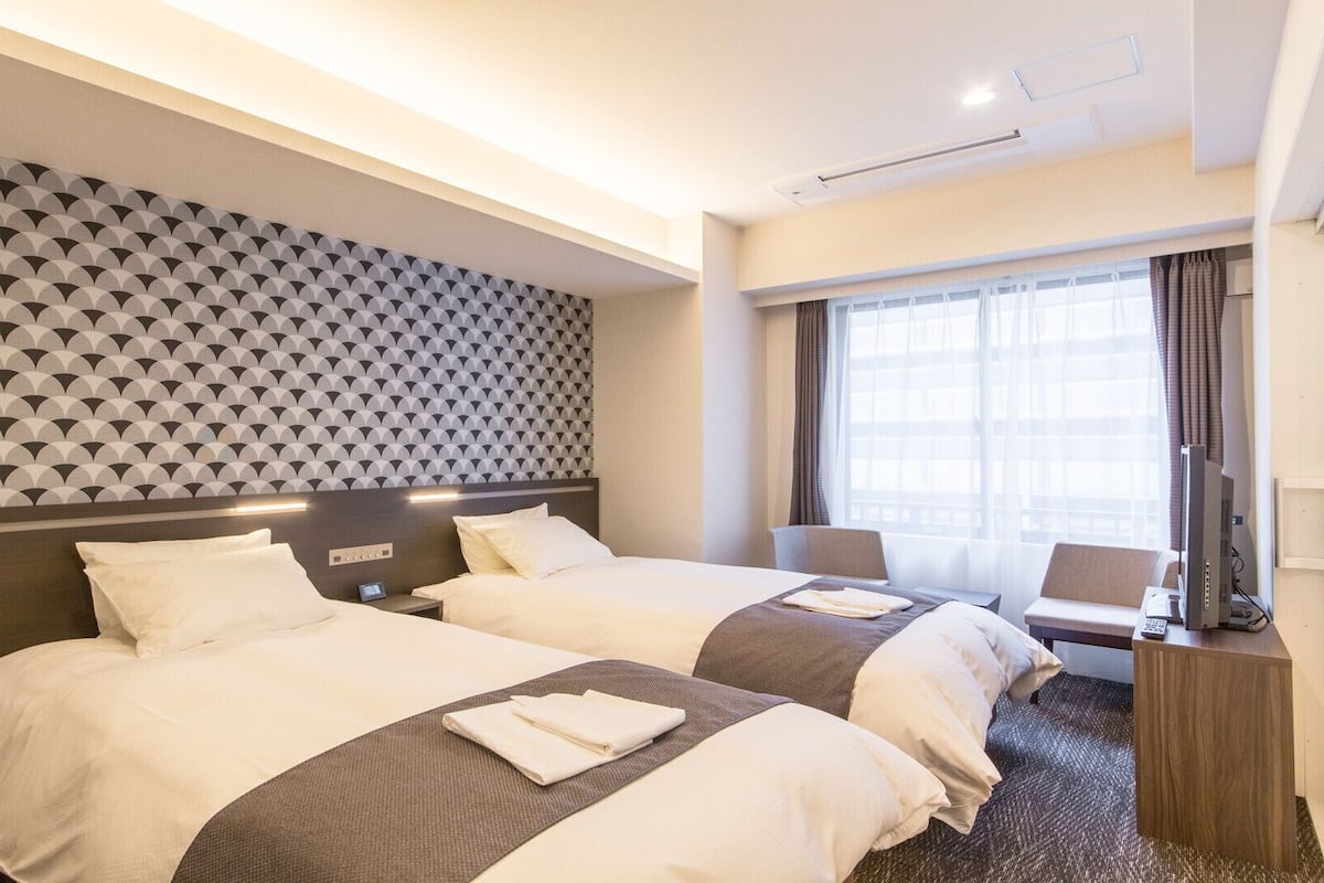 A hotel room with two beds, a patterned wall, a window with curtains, and a desk with a computer.