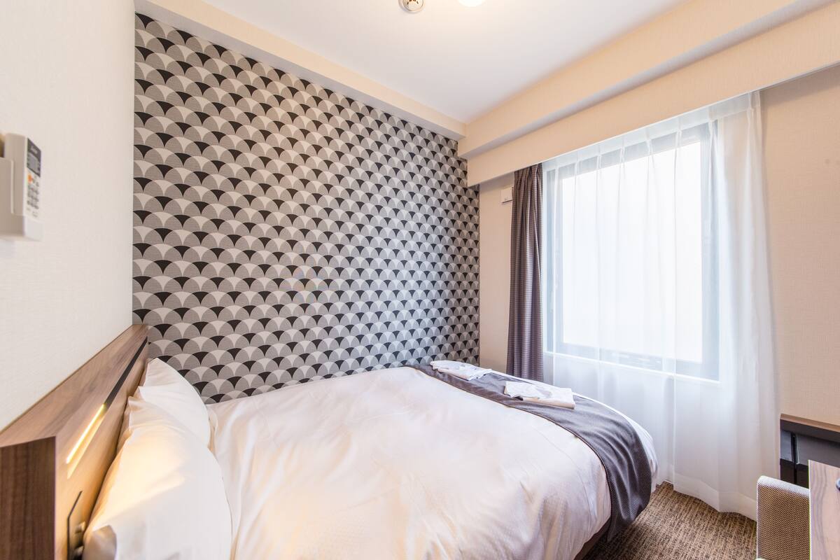 A hotel room with a bed, a headboard with a geometric pattern, a window with curtains, and a small table with a chair.