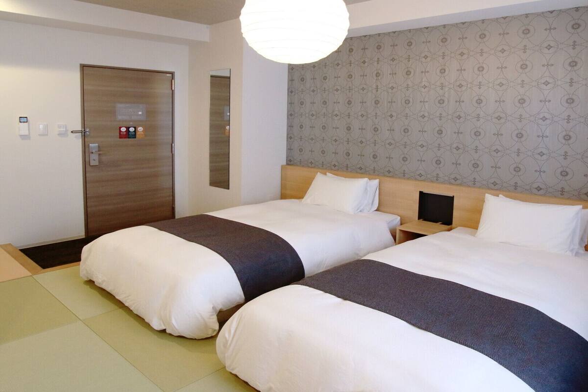 A hotel room with two beds, a wooden door, a mirror, a patterned wallpaper, and a light fixture.