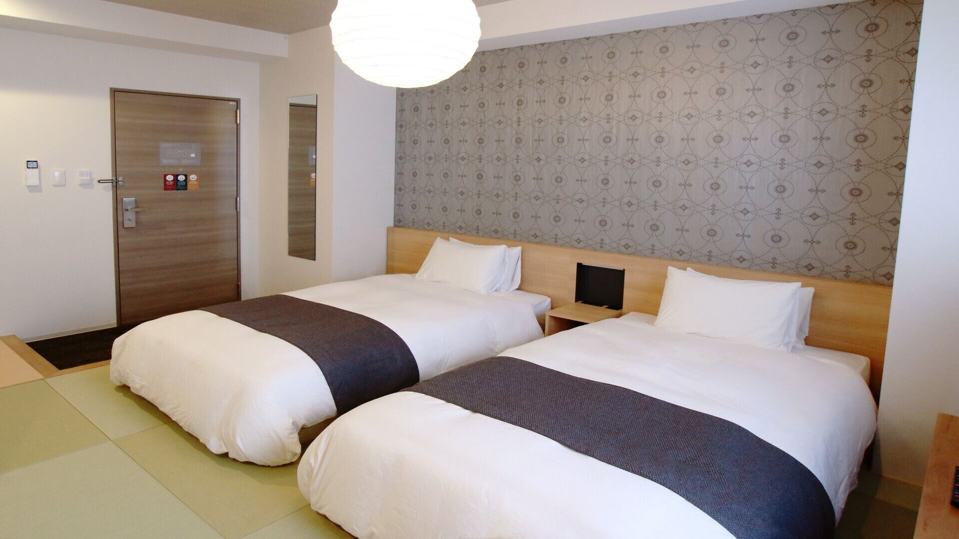 Habitación de hotel con dos camas, una puerta de madera, un espejo y papel tapiz estampado.