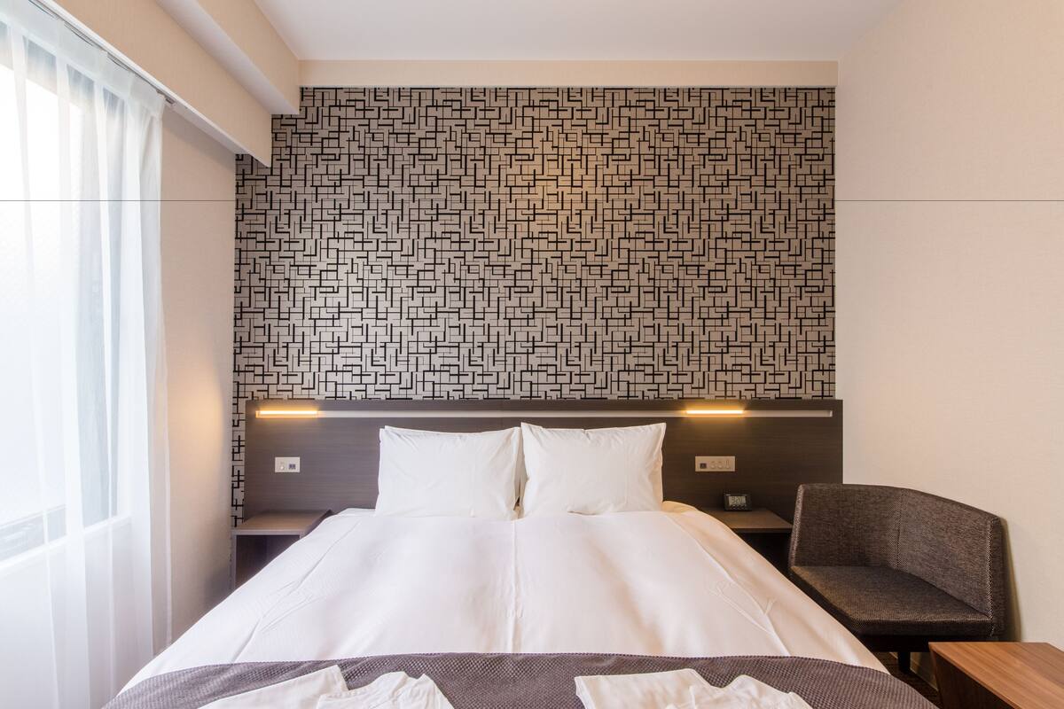 A hotel room with a neatly made bed, a headboard with a geometric pattern, a bedside table with a lamp, and a chair.