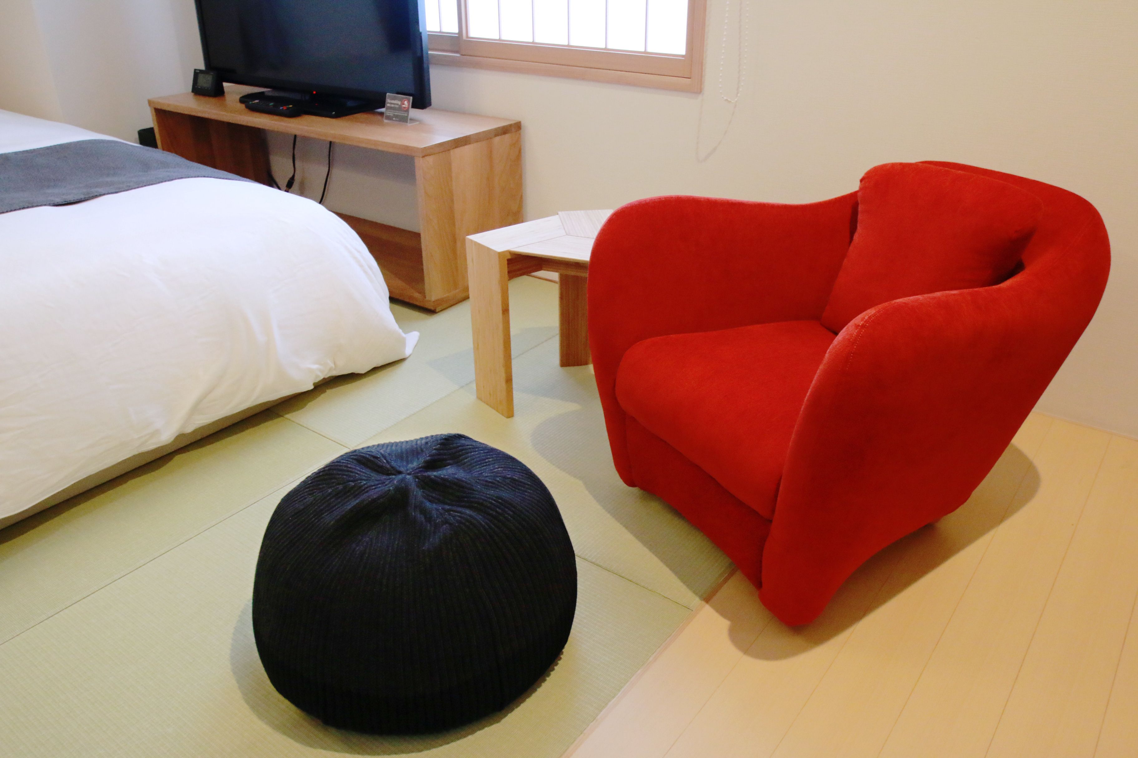 A room with a bed, a red chair, a small table, and a black ottoman.