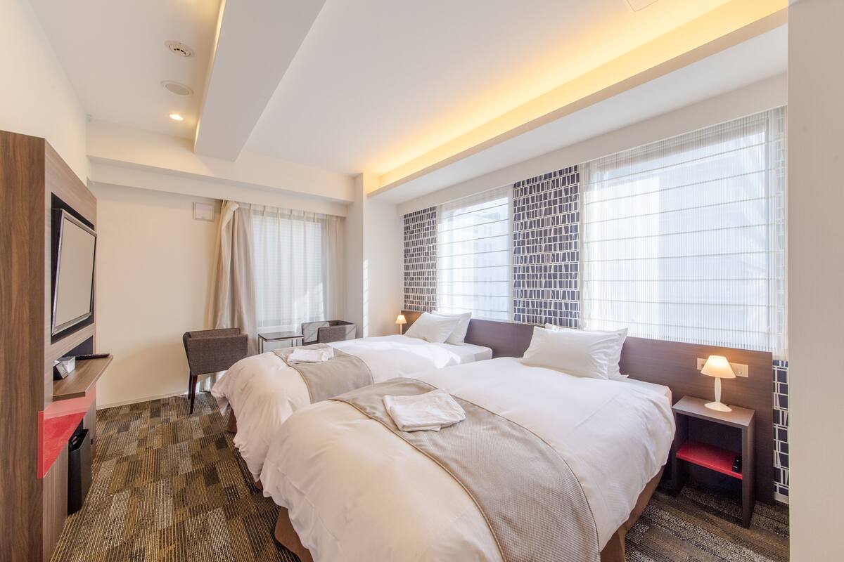 A hotel room with two beds, a TV, a desk, and a window with a view.
