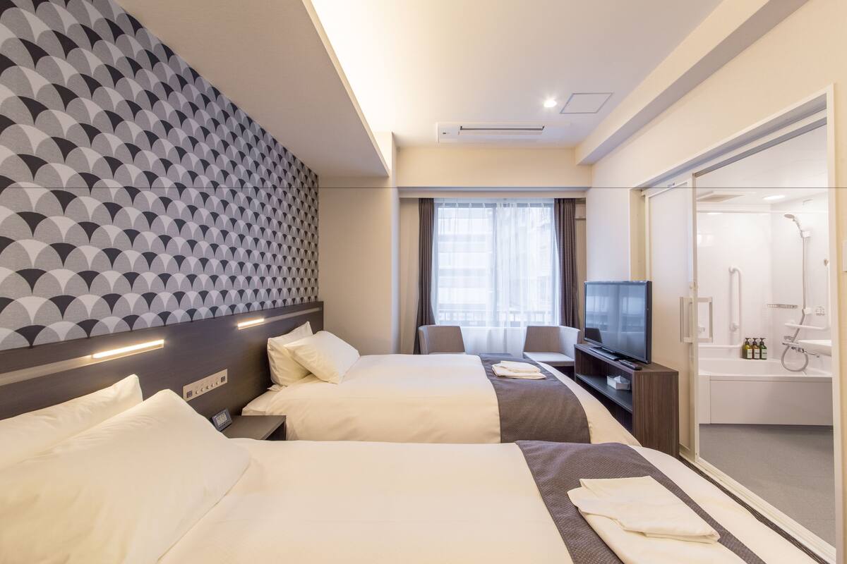 A hotel room with two beds, a TV, a desk with a chair, and a bathroom with a shower and toilet visible through a glass door.